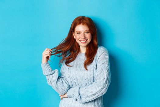 Flirty young woman with red hair, playing with hair and smiling, standing in sweater against blue background