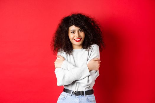Tender young woman hugging herself, feeling comfortable and happy, smiling silly at camera, standing against red background