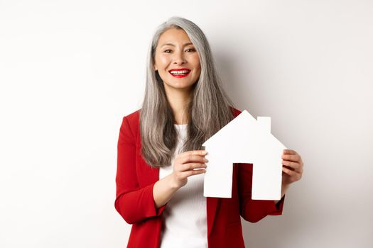 Asian female real estate agent showing paper house cutout, broker smiling friendly and selling property, standing over white background