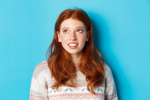 Close-up of dreamy teen girl with red hair, looking at upper left corner and smiling, standing against blue background
