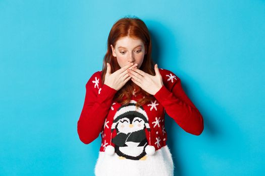 Winter holidays and Christmas Eve concept. Surprised redhead girl gasping, looking down with awe, staring at logo, standing in xmas sweater against blue background