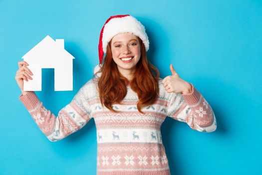 Holiday promos and real estate concept. Satisfied female model with red wavy hair, wearing santa hat and sweater, showing paper house model and thumbs-up, blue background.