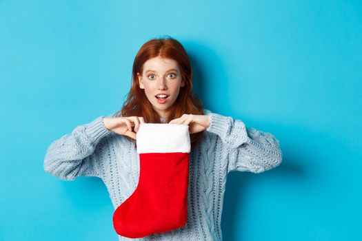 Winter holidays and gifts concept. Funny redhead girl looking surprised, open Christmas stocking and smiling, receiving xmas present, standing against blue background