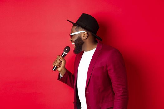 Passionate black male singer performing against red background, singing into microphone, wearing party outfit, standing over red background