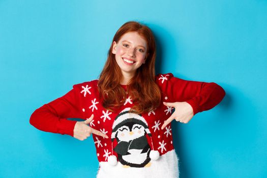 Winter holidays and Christmas Eve concept. Pretty redhead girl pointing fingers at cute xmas sweater with penguin, standing over blue background