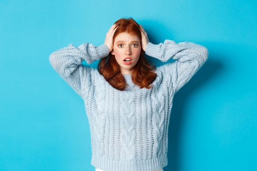 Nervous redhead girl standing overwhelmed, holding hands on head in panic and staring at camera, standing anxious against blue background