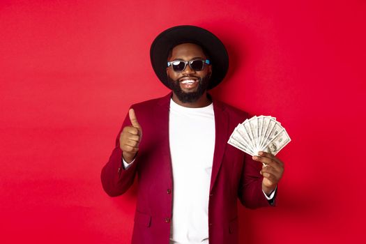 Handsome modern african american man in sunglasses and party clothes, showing thumb up with dollars, earn money and looking satisfied, red background