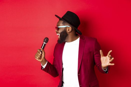Passionate black male singer performing against red background, singing into microphone, wearing party outfit, standing over red background
