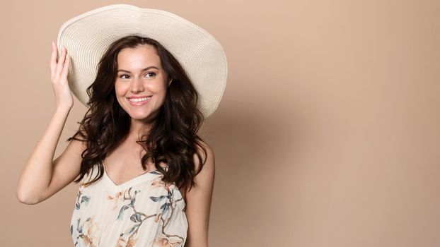 Happy caucasian young woman in summer dress and straw hat looks at empty space on beige studio background. Cheerful lady advertising your summer product