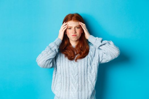 Distressed teenage redhead girl touching head, looking with troubled face expression, standing against blue background, have problem