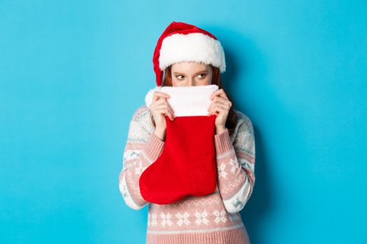 Cute girl cover face with christmas stocking, staring right with cunning gaze, standing in Santa hat and celebrating winter holidays, blue background