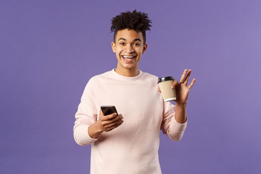Portrait of cheerful young male student with cup of coffee and mobile phone, waving hand to say hi, smiling friendly as greeting person, meet classmate near university campus, purple background