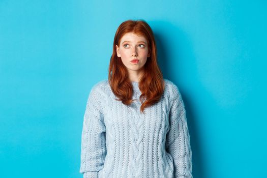 Dreamy redhead girl thinking or making decision, looking at upper left corner logo, standing against blue background