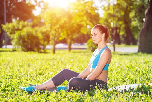 Beautiful smiling girl in sportswear relax in park. Young woman with sitting on green grass after training. Meditation outdoor at sunny summer day. Morning exercises and healthy lifestyle.