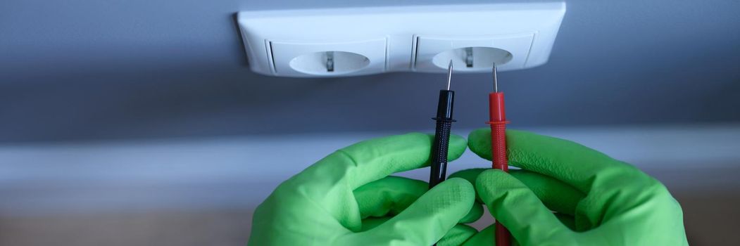 Craftsman in rubber gloves holding tester near electrical outlet at home closeup