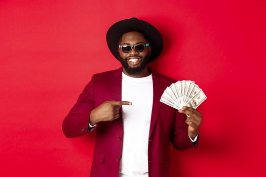 Handsome and stylish african american male model showing money and smiling, wearing sunglasses and fancy hat, standing over red background