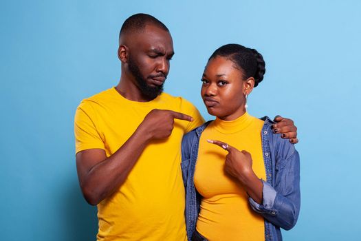African american people pointing index fingers at each other in studio. Playful couple showing sideways directions and sharing embrace in front of camera. Romantic partners in relationship