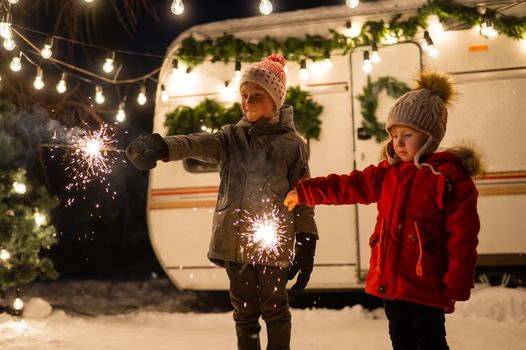 Caucasian red-haired boys hold sparklers by the trailer. Two brothers are celebrating Christmas on a trip.