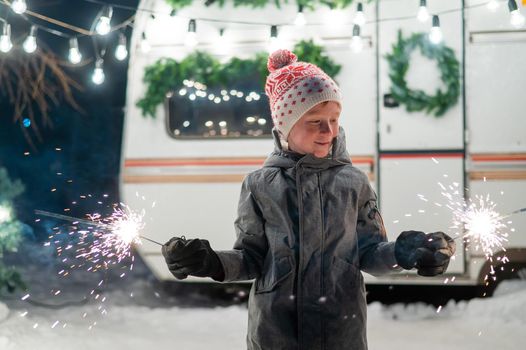 Caucasian red-haired boy holding sparklers by the trailer. Schoolboy celebrates Christmas on a trip.