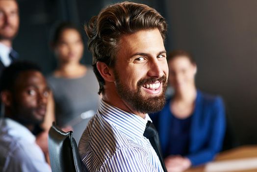 He leads his team through positivity. Portrait of a smiling businessman sitting in an office with colleagues in the background.
