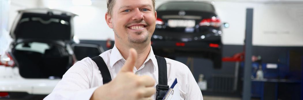 Portrait of a young smiling car mechanic holding thumbs up in car repair shop