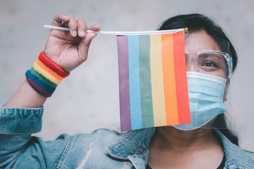Asian lady wearing mask and face shield for protect covid-19 virus holding rainbow flag, symbol of LGBT pride month celebrate annual in June social of gay, lesbian, bisexual, transgender, human rights.