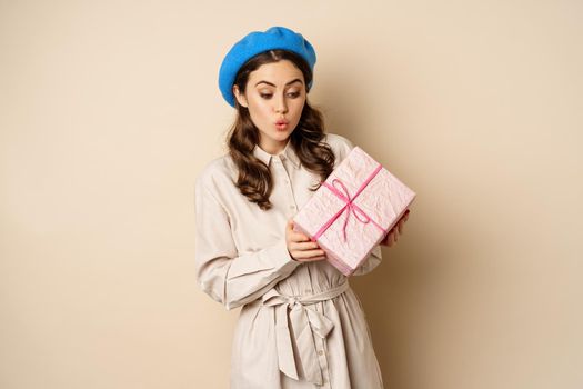 Holidays and gifts concept. Beautiful girl receive gift box and looking happy, holding pink wrapped present with joyful face expression, beige background
