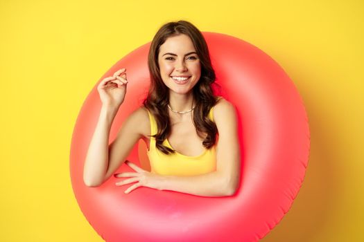 Close up portrait of beautiful brunette woman model inside pink swimming ring, beach accessories and summer vacation concept, yellow background