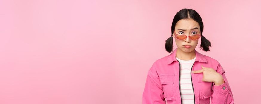 Portrait of asian girl looks confused and points at herself, perplexed face, stares with disbelief at camera, stands over pink background
