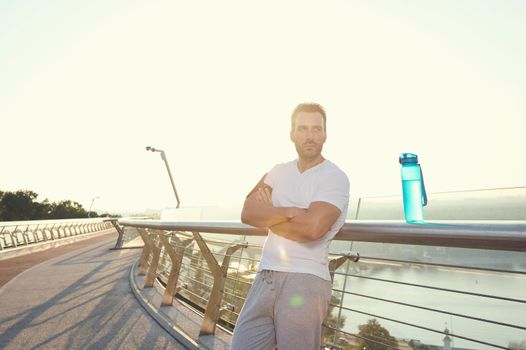 Close-up portrait of an attractive middle aged muscular build Caucasian man in sportswear standing on city bridge and relaxing after heavy workout outdoor. Sunbeams falling on the treadmill at dawn