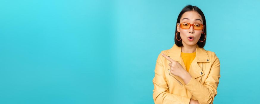 Image of asian woman looks intrigued, asks question about item or store, points finger left with surprised face expression, stands over blue background
