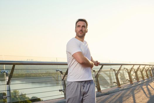 Middle aged European man in sportswear exercising outdoor on the city bridge at dawn on a warm sunny summer day. Sport, active and healthy lifestyle concept. Keep you body fit