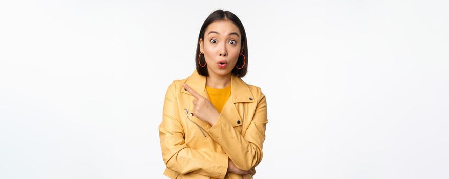 Portrait of beautiful asian woman express intereest and excitement about advertisement, store discount, pointing finger left, looking amazed, standing over white background