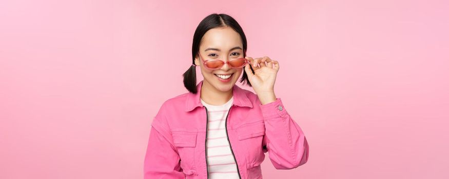 Eyewear advertisement. Stylish modern asian girl touches sunglasses, wears pink, poses against studio background. Copy space
