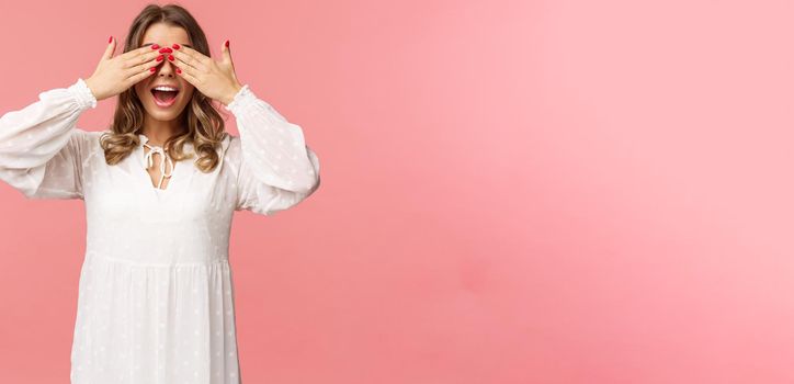 Excited cute blond girl in white spring dress waiting for gifts at surprise party, cover eyes blindfolded standing pink background, open mouth in amazement and anticipation