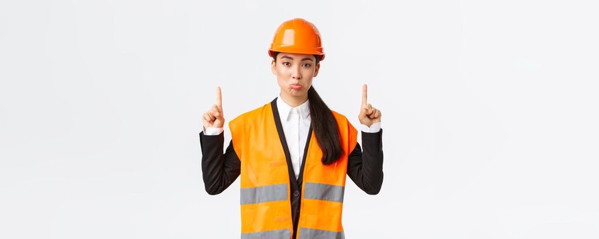 Building, construction and industrial concept. Displeased sad asian female architect complaining, wearing safety helmet and reflective clothing, pouting upset as pointing fingers up