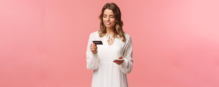 Portrait of attractive and cute blond girl with short curly hairstyle, white dress, holding smartphone and credit card, insert digit numbers to register in shopping app, buy online, pink background