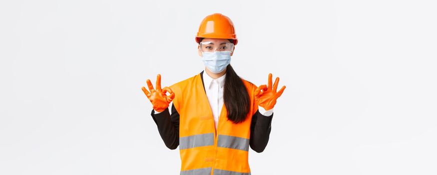 Covid-19 safety protocol at enterpise, construction and preventing virus concept. Satisfied asian female engineer, industrial woman face mask showing okay gesture, well done, praising good work