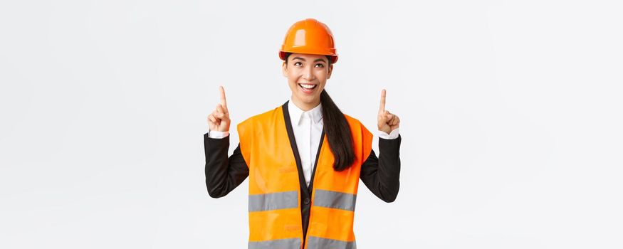 Building, construction and industrial concept. Successful pleased asian female engineer in safety helmet inspect object, looking and pointing fingers up satisfied. Architect pleased with good result