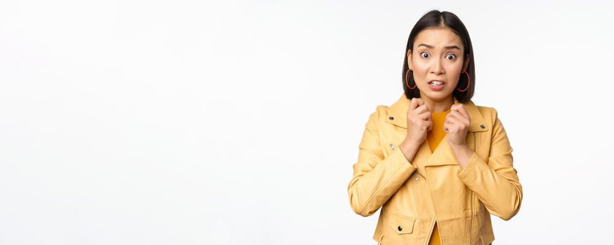 Image of scared asian woman looking insecure and worried at camera, gasping shocked, standing worried against white background