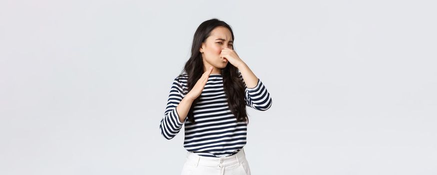 Lifestyle, beauty and fashion, people emotions concept. Disgusted korean girl shut nose and waving hand from disgust and aversion as sniff awful disgusting smell, standing white background