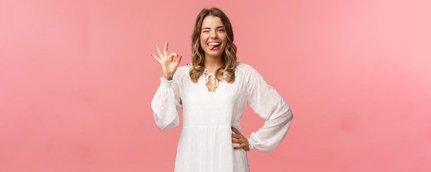 Portrait of happy smiling blond woman with short curly haircut, show tongue and okay sign, say alright, agree or approve, rate great product, guarantee quality, stand pink background