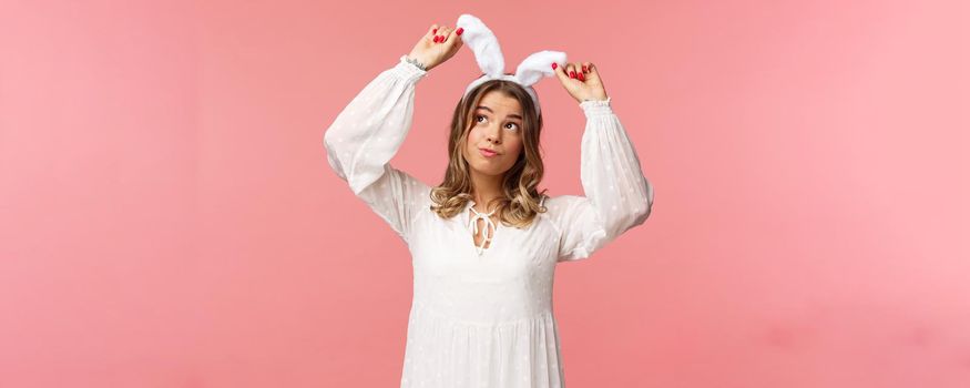Holidays, spring and party concept. Portrait of silly cute blond girl in rabbit ears, looking up daydreaming, smiling lovely wear white dress, standing over pink background carefree
