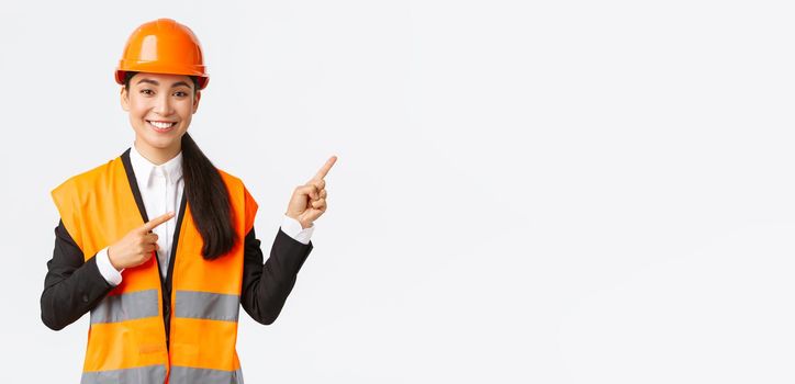 Building, construction and industrial concept. Smiling female asian engineer in safety helmet and reflective clothing showing object, pointing fingers right, saleswoman introduce estate to client