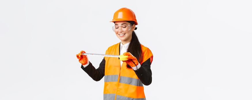 Pleased smiling asian female architect achieve good results, looking satisfied at tape measure after measuring layout at construction area, standing in safety helmet and reflective clothing