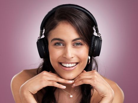 Where words fail music speaks. Shot of a beautiful young smiling woman with her hands rested on her chin while wearing headphones against a pink background.