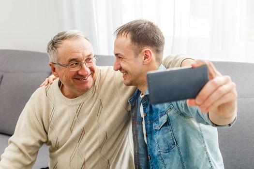 Happy moment. Cheerful young man taking a selfie with his upbeat elderly father waving at the camera and smiling pleasantly