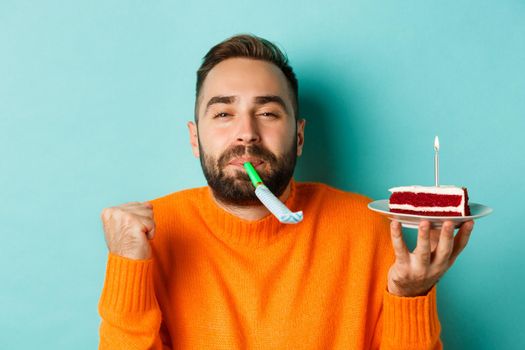 Close-up of funny adult man celebrating his birthday, holding bday cake with candle, blowing party wistle and rejoicing, standing over light blue background