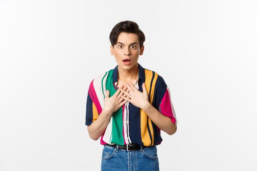 Portrait of surprised gay man holding hands on heart, looking with disbelief and amazement at camera, standing over white background