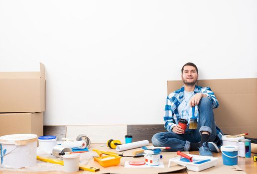 Happy smiling guy relaxing on floor with coffee after moving in new house. House remodeling and interior renovation concept. Young bearded man sitting on floor among cardboard boxes and painting tools